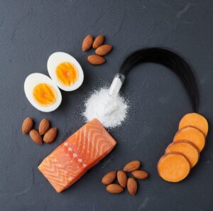 Overhead flat-lay of biotin supplement capsules alongside natural food sources like eggs, salmon, and almonds, with a strand of hair connecting them.